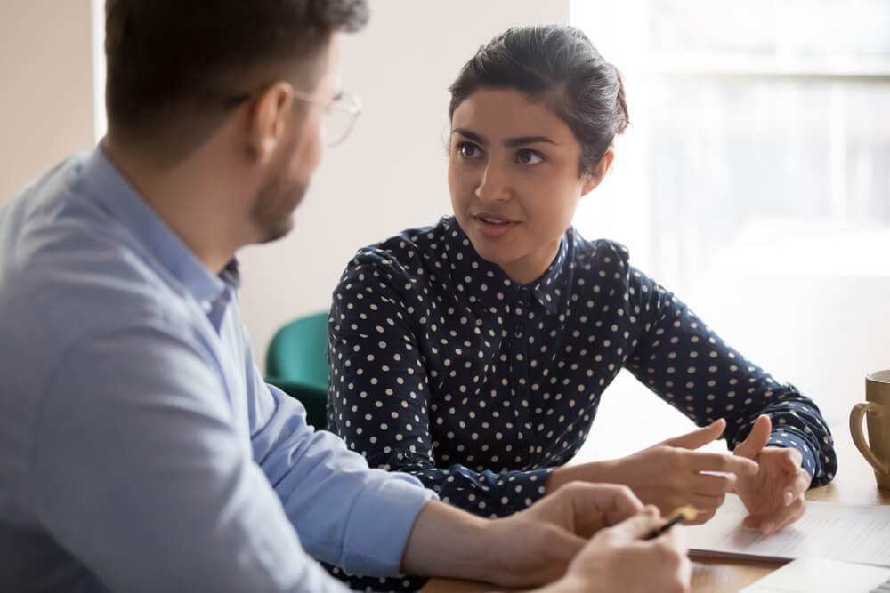 A woman in a polka dot shirt is engaged in conversation with a man in glasses and a light blue shirt. In a bright room with large windows, they sit at a table adorned with papers and a mug, possibly discussing insights related to the public sector.